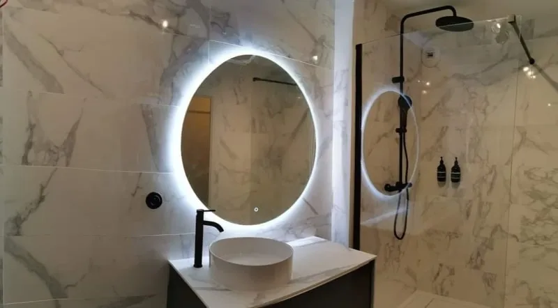 Détail de salle de bains : vasque posée, mitigeur noir et miroir rond rétroéclairé sur carrelage effet marbre
